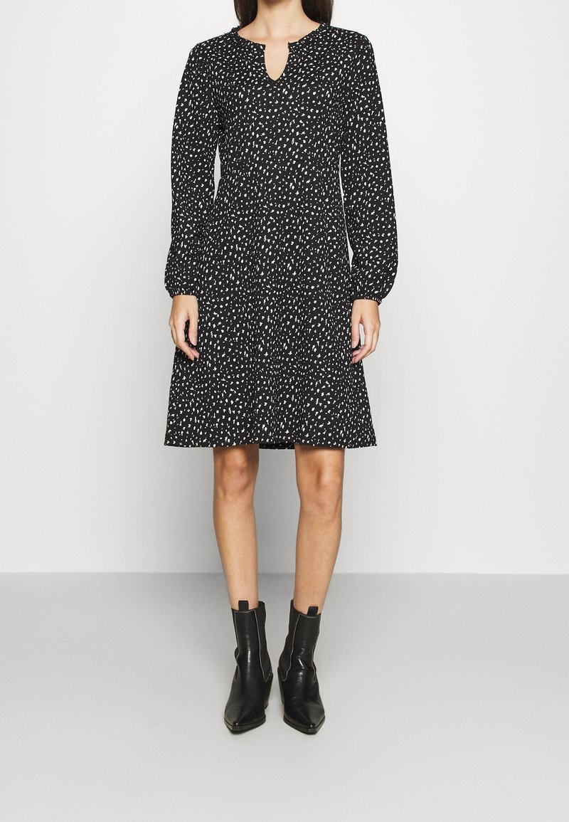 Femme portant une robe noire à manches longues avec des taches blanches, un petit décolleté en V, un ourlet à hauteur des genoux, et des bottines noires.