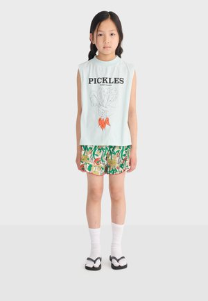 Jeune fille aux cheveux tressés portant un débardeur "PICKLES", un short floral coloré, des chaussettes blanches et des sandales noires, debout devant un fond uni.