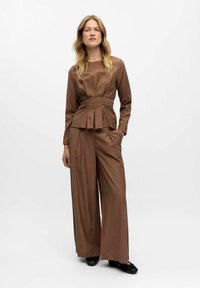 Femme debout avec une main dans la poche, portant un haut plissé à manches longues marron et un pantalon large avec des chaussures noires sur fond blanc.