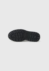 Suela de zapato de goma negra con patrones de textura, con profundos surcos y crestas para tracción. Tacón plano, detalle de marca mínimo.