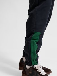 Dolne części nóg w czarnych spodniach z zielonymi paskami po bokach oraz brązowych tenisówkach z białymi sznurówkami i skarpetkami na jasnym tle.