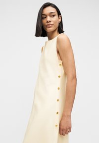 Γυναίκα με ίσια σκούρα μαλλιά που φοράει αμάνικο κρεμ φόρεμα με χρυσά κουμπιά κατά μήκος της πλευράς, κοιτάζοντας προς τα αριστερά.