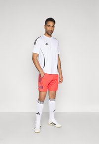 Biele športové tričko s čiernymi doplnkami, jasne oranžové šortky s odznakom, biele ponožky s čiernymi pruhmi a kopačky s čiernymi detailmi.