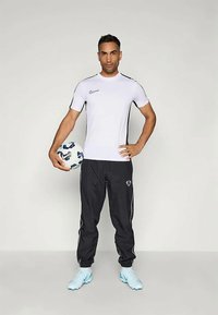 Valkoinen urheilupaita mustilla yksityiskohdilla, mustat juoksuhousut ja vaaleansiniset jalkapallokengät. Pitelee kuvioitua jalkapalloa.