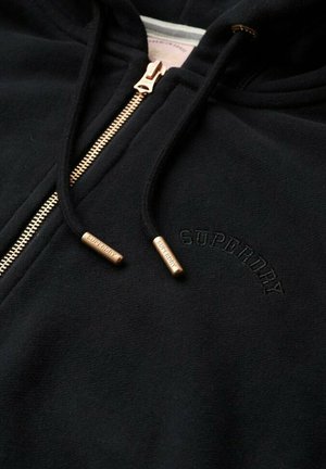 Sudadera negra con cremallera y detalles metálicos en dorado, con un logo bordado de "Superdry" en negro tonal en el pecho. Textura suave.