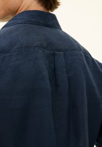 Camicia in lino blu navy con colletto, dettaglio del pleat sul retro e tessuto testurizzato. L'immagine mostra il capo da una prospettiva posteriore.