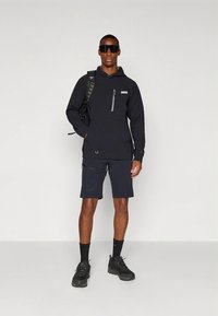 Sweatshirt preta com bolso frontal e fecho de correr cinzento, combinada com calções pretos. Modelo usa óculos de sol e sapatilhas pretas de caminhada, posicionado contra um fundo branco.