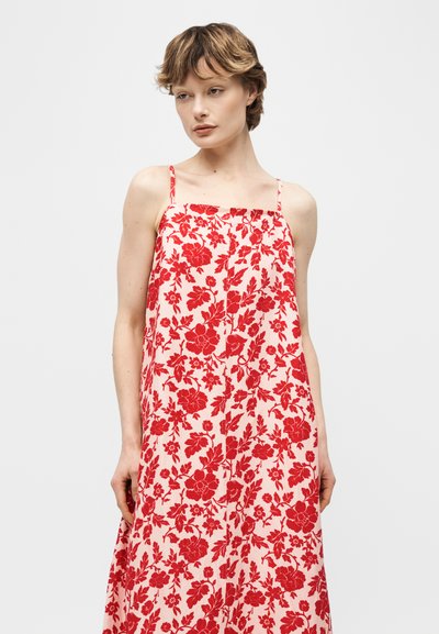 Γυναίκα που φοράει αμάνικο κόκκινο φόρεμα με λουλούδια και λεπτές τιράντες, μπροστά σε απλό ανοιχτό φόντο.