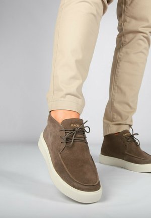 Scarpe in suede marrone con finitura liscia, suola in gomma bianca e design con lacci. Abbinamento con pantaloni beige chiaro per un look casual.