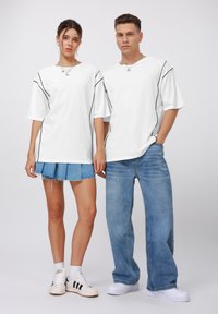 White t-shirts with black side seams, paired with a denim skirt and loose jeans. Both models wear sneakers and silver chain necklaces.