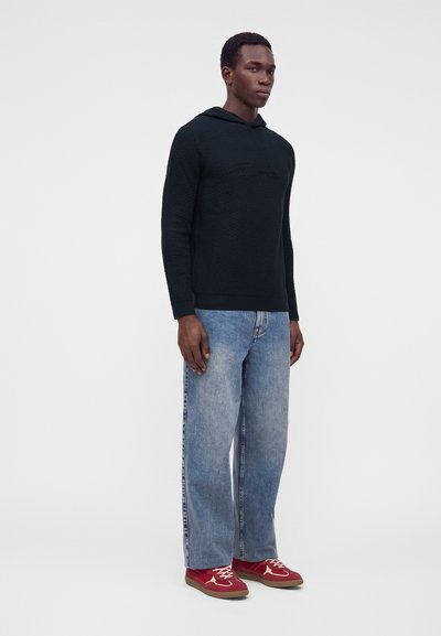 Homme portant un sweat à capuche noir texturé, un jean large bleu clair et des baskets rouges, debout devant un fond clair uni.