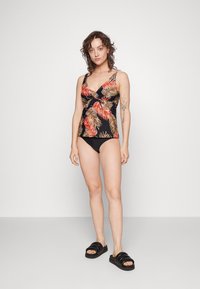 Schwarzer Tankini mit rotem und goldenen Palmblattmuster; V-Ausschnitt und geraffte Taille. Kombiniert mit schwarzen Bikini-Höschen und klobigen schwarzen Sandalen.