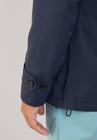 Giacca della marina con una texture liscia, dotata di due bottoni neri sui polsini e di un cordino sul fondo, indossata sopra pantaloni azzurri chiari.