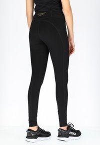 Zwarte leggings met een hoge tailleband, voorzien van textuurstiksels en logo-detail. Draagt met zwarte sportschoenen met witte accenten.