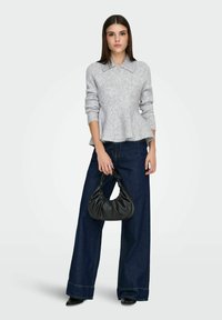 Světle šedý peplum svetr z měkké látky, kombinovaný s tmavě modrými džínami s širokými nohavicemi. Model drží černou, nařasenou kabelku.