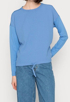 Frau trägt ein hellblaues Langarmshirt mit Kordelzugsaum und blaue Jeans, steht mit einer Hand in der Hüfte vor weißem Hintergrund.