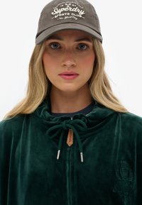 Grön sammetszip-up hoodie med dragsnöre, med en broderad logotyp. En grå keps med vit text bärs på huvudet.