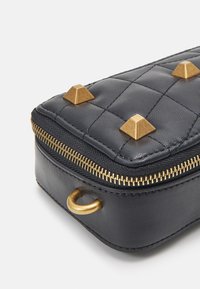 Černá prošívaná kožená taška se zlatými pyramidovými hroty, vybavená zlatým zipem a kulatým kovovým detailem po straně.