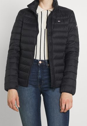 Woman wearing black quilted zip-up jacket over white collared shirt with black vertical stripes and blue jeans, standing against plain backdrop.