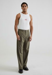 Hvid tanktop med rød tekst "PROHIBITED SOCIAL CLUB" kombineret med højtaljede olivengrønne bukser og sorte sko, sat mod en neutral baggrund.