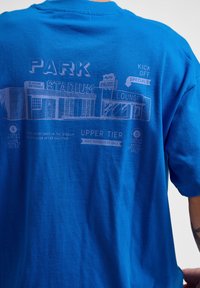 Blå t-shirt gjord av bomull, med ett tryckt mönster av en stadion med texten "PARK", "STADIUM" och lounge-detaljer i ljusare blått.