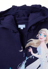 Hoodie zippé bleu marine avec un design imprimé d'un personnage aux cheveux blonds, portant une robe ornée de cristaux et d'accents.