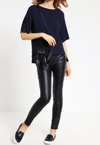 Nainen, jolla on päällä tummansininen ribbipintaista lyhythihaista toppia, mustat nahkahousut, musta olkahihnallinen laukku, jossa on tupsuja, ja mustat liukuvaljaat valkoisilla pohjilla.