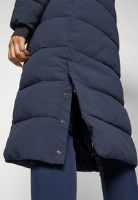 Mørkeblå pufferjakke med quiltet polstring, med en sidesnap-placket. Myk, teksturert stoff med et knelengde-design.