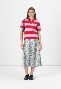 Le modèle se tient devant un fond blanc uni, portant un polo rayé rouge et rose, une jupe argentée brillante et des chaussures noires.