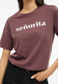 Bordowa bawełniana koszulka z krótkimi rękawami z białym nadrukiem słowa "señorita". Dół koszulki jest krótki, zestawiona z czarnymi jeansami.