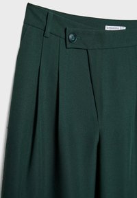 Dunkelgrüne, taillierte Hose mit vorderen Falten, einem einzelnen Knopfverschluss und einem glatten, strukturierten Stoff. Etikett im Bund sichtbar.