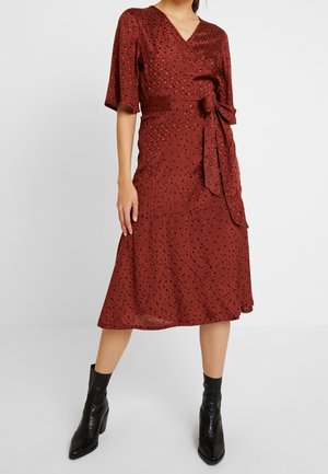 Kobieta w rdzawo-czerwonej sukience do kolan w czarne grochy, z przewiązaniem w pasie i czarnymi botkami, stojąca na jasnym tle.