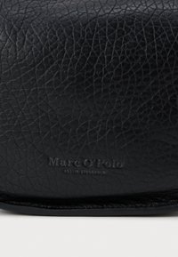 Sac en cuir noir avec une surface texturée. Présente le nom de la marque embossé, "Marc O'Polo." Design simple, bords arrondis et finition douce.