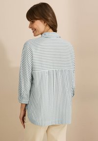 Camicia a righe blu chiaro con piega centrale sul retro, maniche a tre quarti, texture morbida e orlo arrotondato. Sottili righe verticali di un blu più scuro.