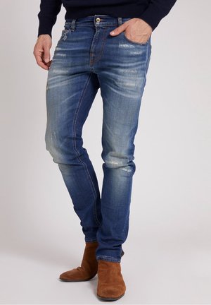 Jeans de mezclilla azul oscuro con corte ajustado, que presentan un ligero desgaste y detalles desgastados. La tela tiene una textura suave con costuras en naranja.