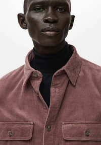 Manchesterskjorta i dammros, med två bröstfickor och knäppning fram. Bärs över en svart polotröja, där det texturerade tyget syns.