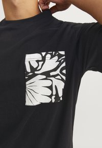 Sort bomulds T-shirt med en hvid lomme og et iøjnefaldende sort botanisk trykdesign. Rund halsudskæring, korte ærmer og almindelig pasform.