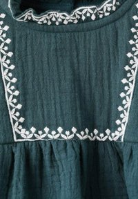 Tirkizna pamučna haljina s bijelim cvjetnim vezom duž izreza i rubova. Tkanina ima teksturirani, naborani izgled.