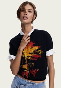 Schwarzes, kurz geschnittenes T-Shirt mit einem lebhaften Grafikmotiv eines gelben und roten Palmenbaums. Kombiniert mit einem weißen Hemd mit Kragen darunter und einer Jeans.