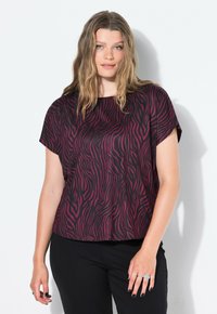 Mujer con cabello rubio ondulado que lleva una blusa de manga corta a rayas de cebra negra y morada oscura y pantalones negros, de pie frente a un fondo blanco.