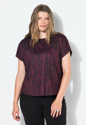 Donna con capelli biondi ondulati, che indossa una blusa a maniche corte zebra a strisce nere e viola scuro e pantaloni neri, in piedi contro uno sfondo bianco.