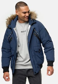 Marineblå isoleret jakke med en faux fur hætte, med flere lynlåslommer og ribbede manchetter. Båret over en grå T-shirt.