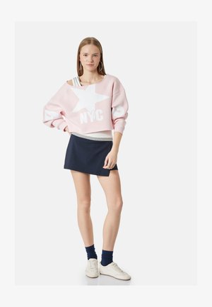 Ung kvinde står med hånden på hoften iført lyserød cropped sweatshirt med hvid stjerne og "NYC", navy nederdel, hvide sneakers og mørke sokker.