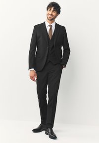 Sort tredelt jakkesæt med en tætsiddende jakke, vest og tilspidsede bukser. Kombinationen omfatter en hvid skjorte og et brunt slips, fuldendt med sorte sko.