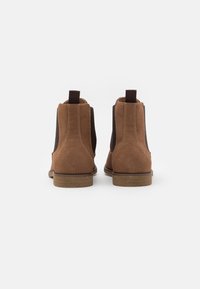 Paire de bottines Chelsea en daim marron avec des panneaux élastiques latéraux foncés et des languettes à tirer, vues de dos sur un fond blanc.