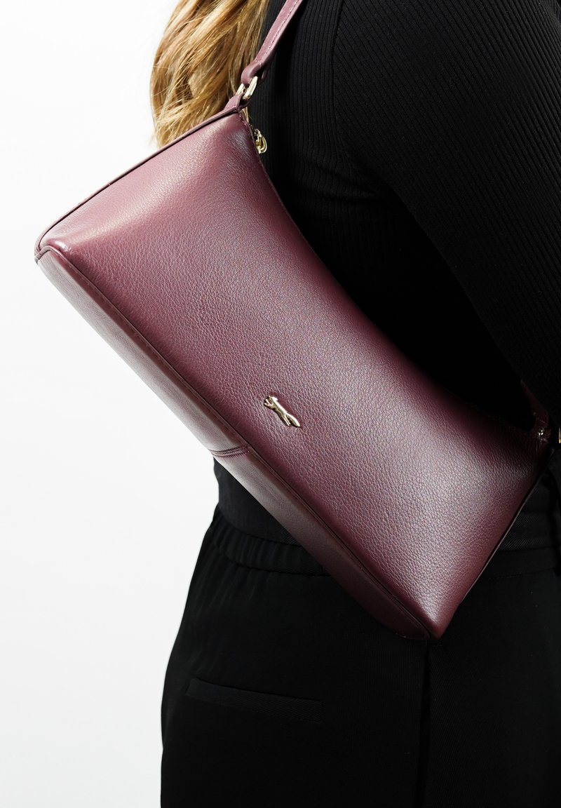 Femme vêtue de noir porte un sac à bandoulière en cuir bordeaux lisse avec un petit logo doré sur le côté.
