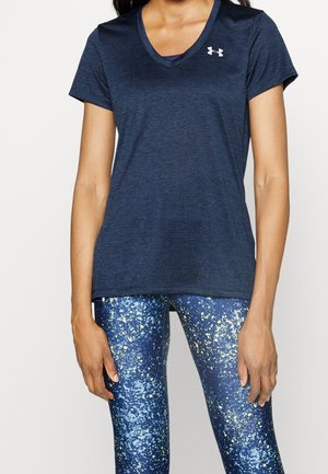 Femme portant un t-shirt Under Armour bleu marine à col en V et des leggings bleus à motif galaxie, debout devant un fond blanc uni.