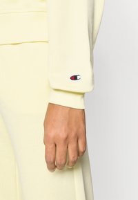 Κίτρινο φούτερ με ριμπ μανσέτα, διακοσμημένο με μικρό κεντητό λογότυπο σε κόκκινο, άσπρο και μπλε  κοντά στον καρπό. Μαλακή υφή.