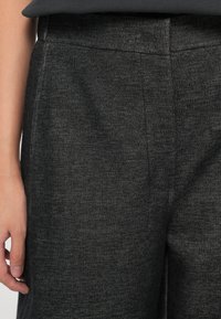 Gros plan sur un pantalon gris foncé texturé porté par une personne avec un haut noir, montrant la taille et une partie de la main gauche.