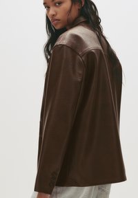 Donna con lunghi capelli scuri che indossa una giacca di pelle marrone scuro oversize e jeans chiari, che guarda oltre la spalla su uno sfondo semplice.